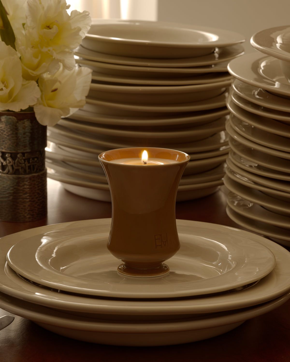 Lit Ambered Apple candle on a plate atop a stack of dinnerware, with white flowers in a vase and sheer curtains behind