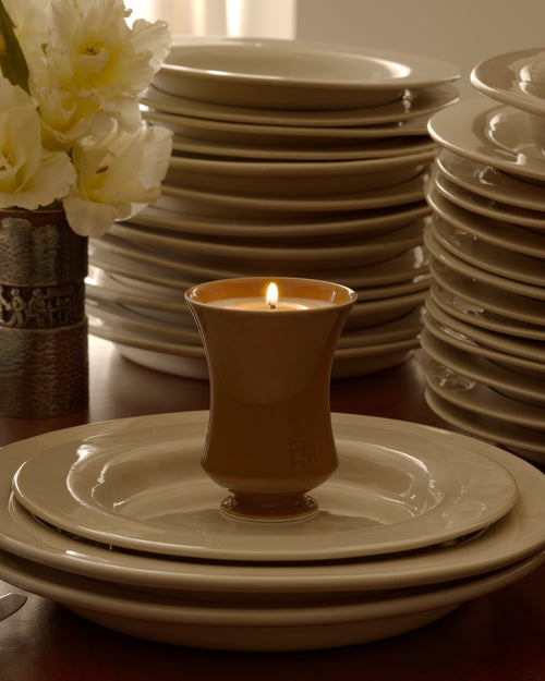 Lit Ambered Apple candle on a plate atop a stack of dinnerware, with white flowers in a vase and sheer curtains behind