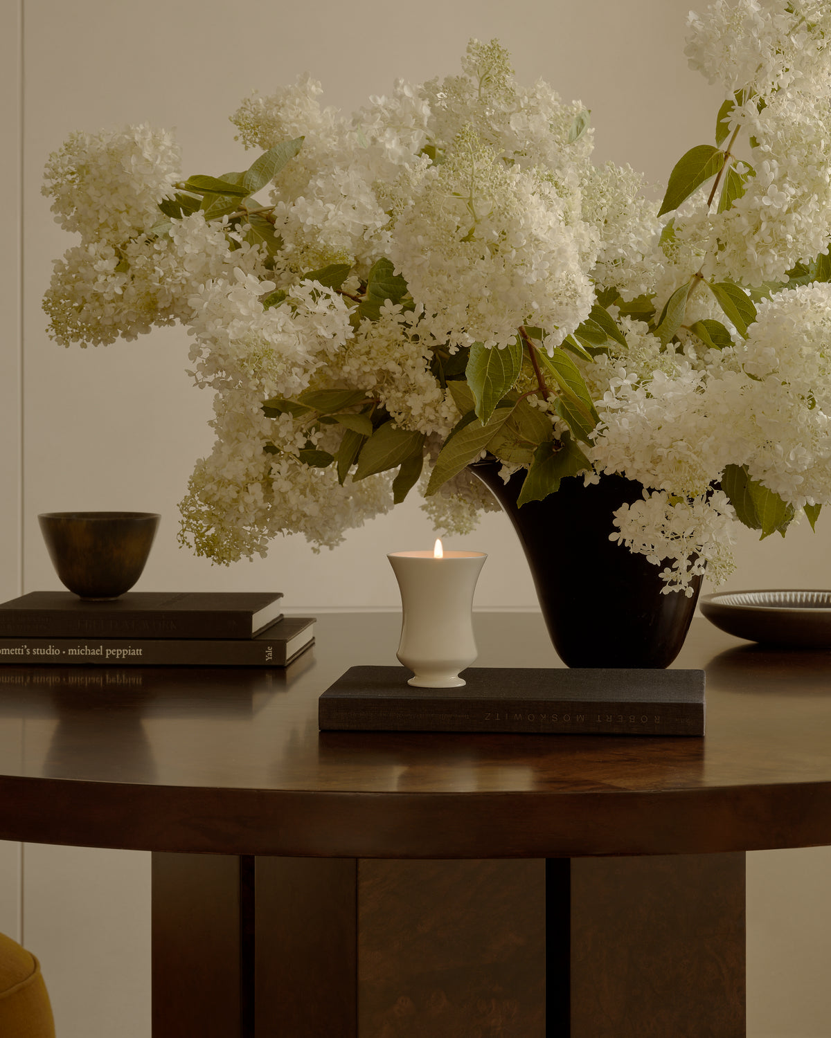 Lit Cashmere Musk candle on a stack of books beside a vase of white hydrangeas